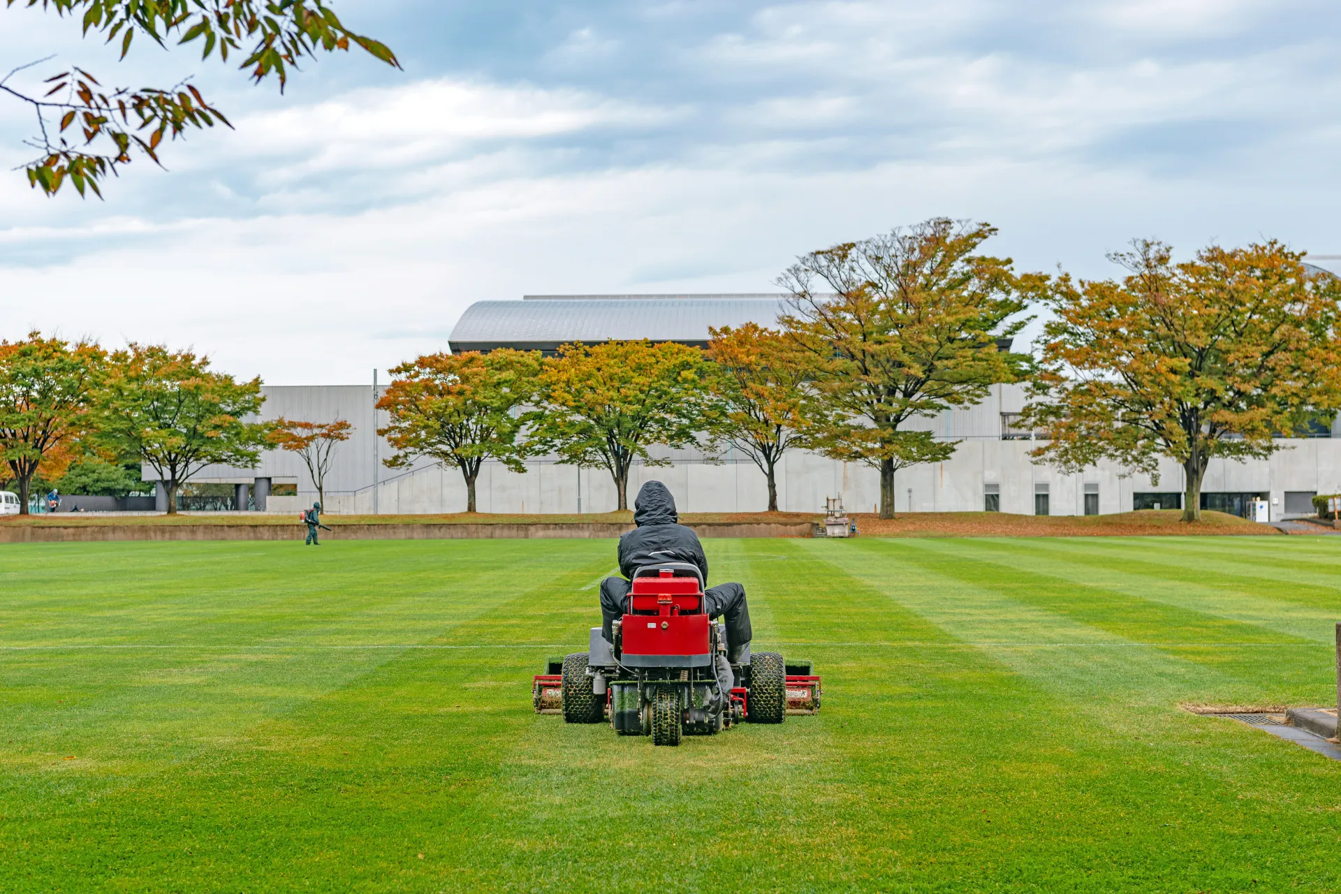 Person rides a red mower across a green field with trees and a white building in the background