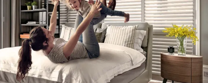 Woman lying on bed joyfully lifts child. Other person partially visible. Bright room.