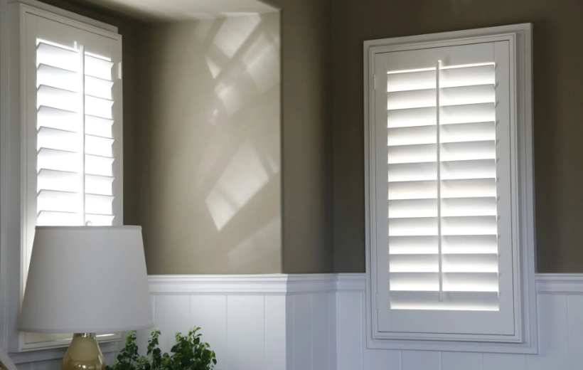 Two white shuttered windows with a lamp and neutral-colored walls.