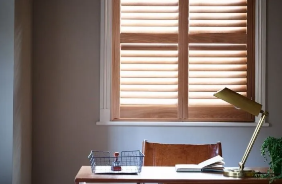 Wooden shutters on a window above a desk with a lamp, chair, and open book.