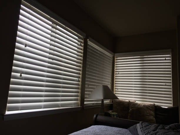 Room with closed blinds casting shadows; bed and lamp visible.