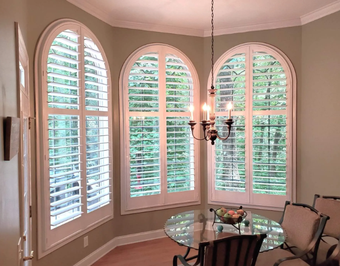 Arched windows with white shutters in a dining room; glass table and chandelier.