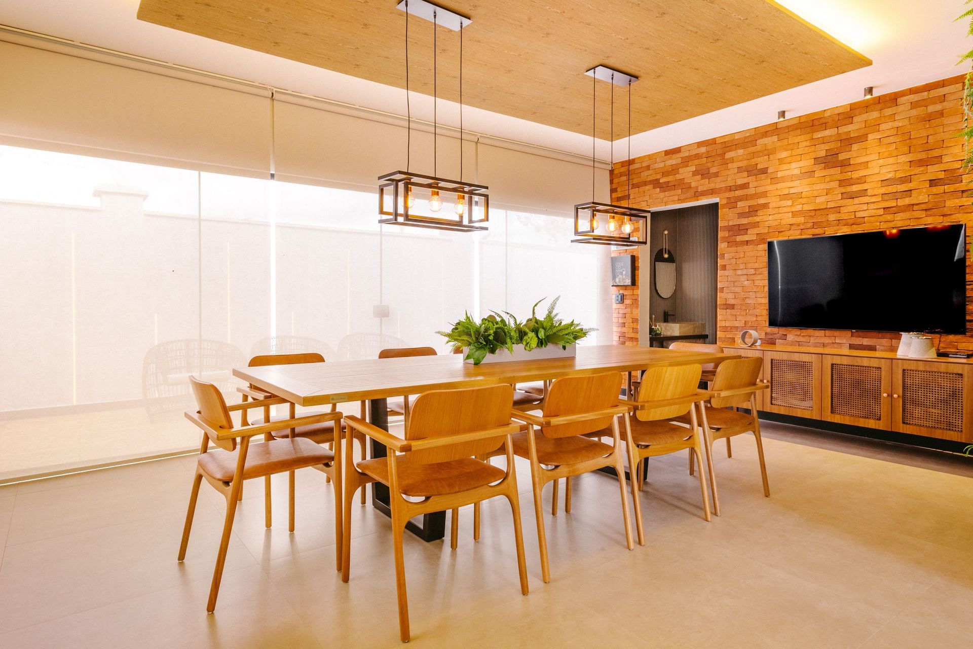 Bright modern dining room with wooden table and chairs, brick wall, pendant lights, and wall-mounted TV