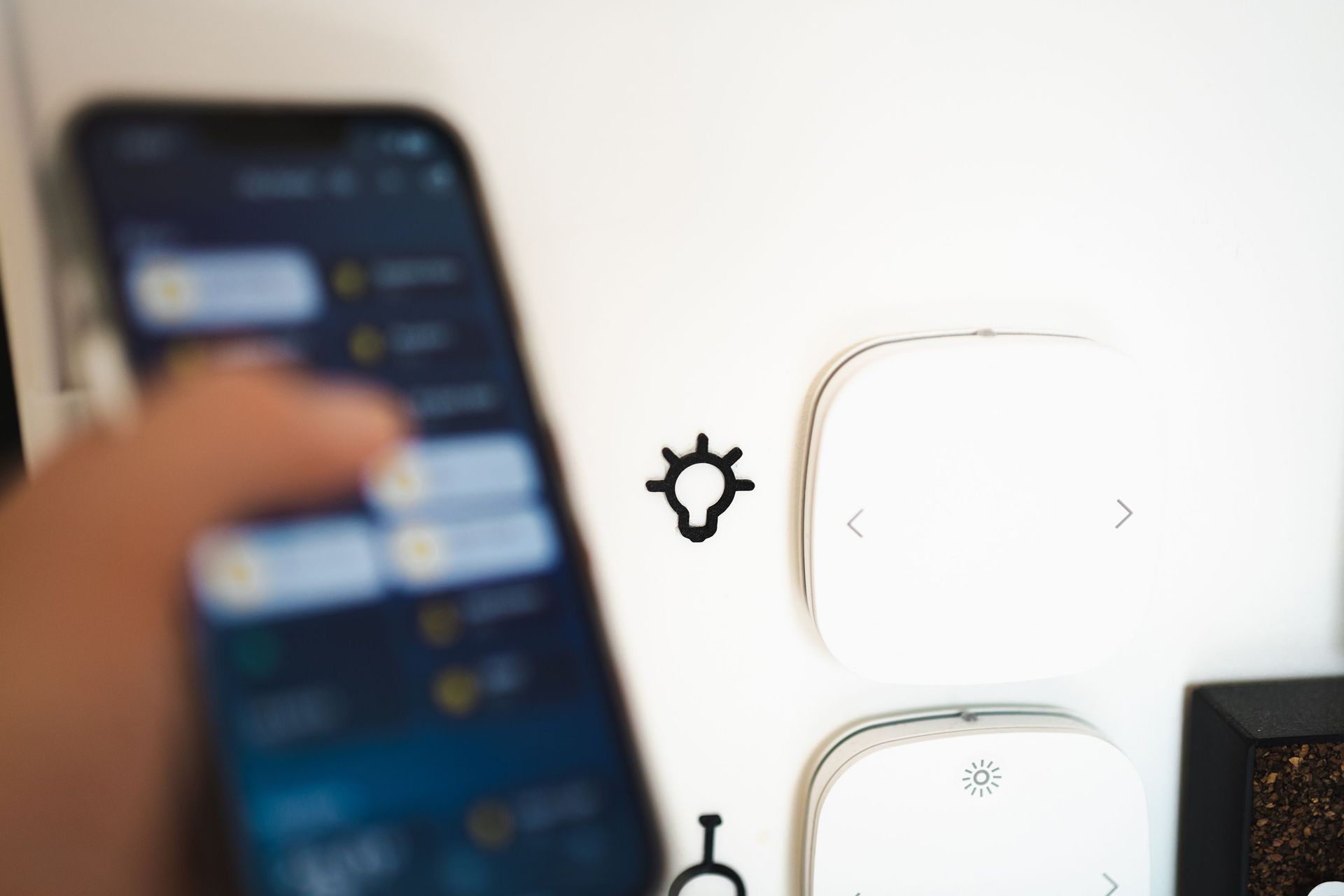 Blurred hand holding smartphone in front of wall-mounted smart switches and a thermostat.
