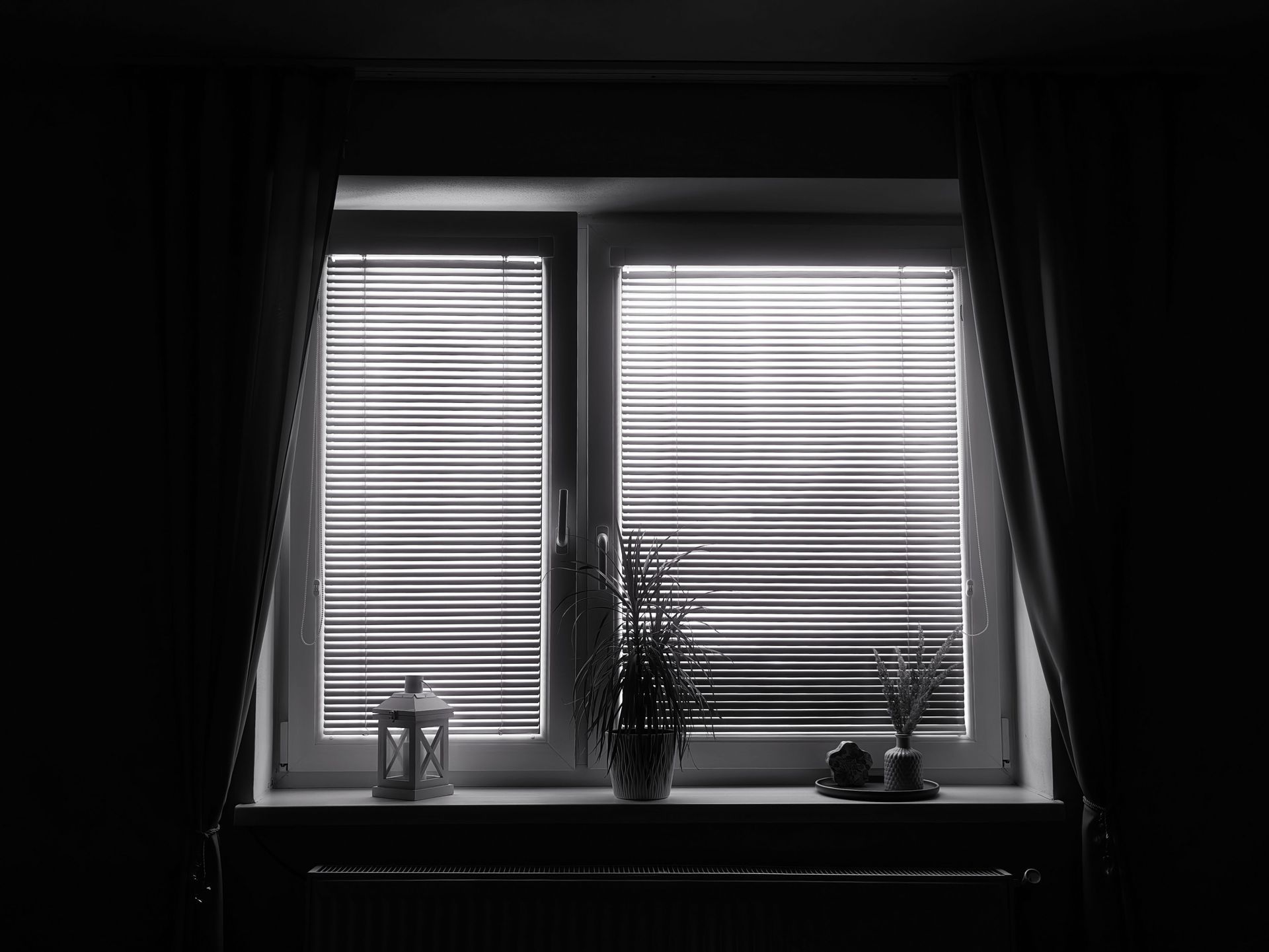 Dim window with closed blinds, framed by dark curtains, and small potted plants on the sill