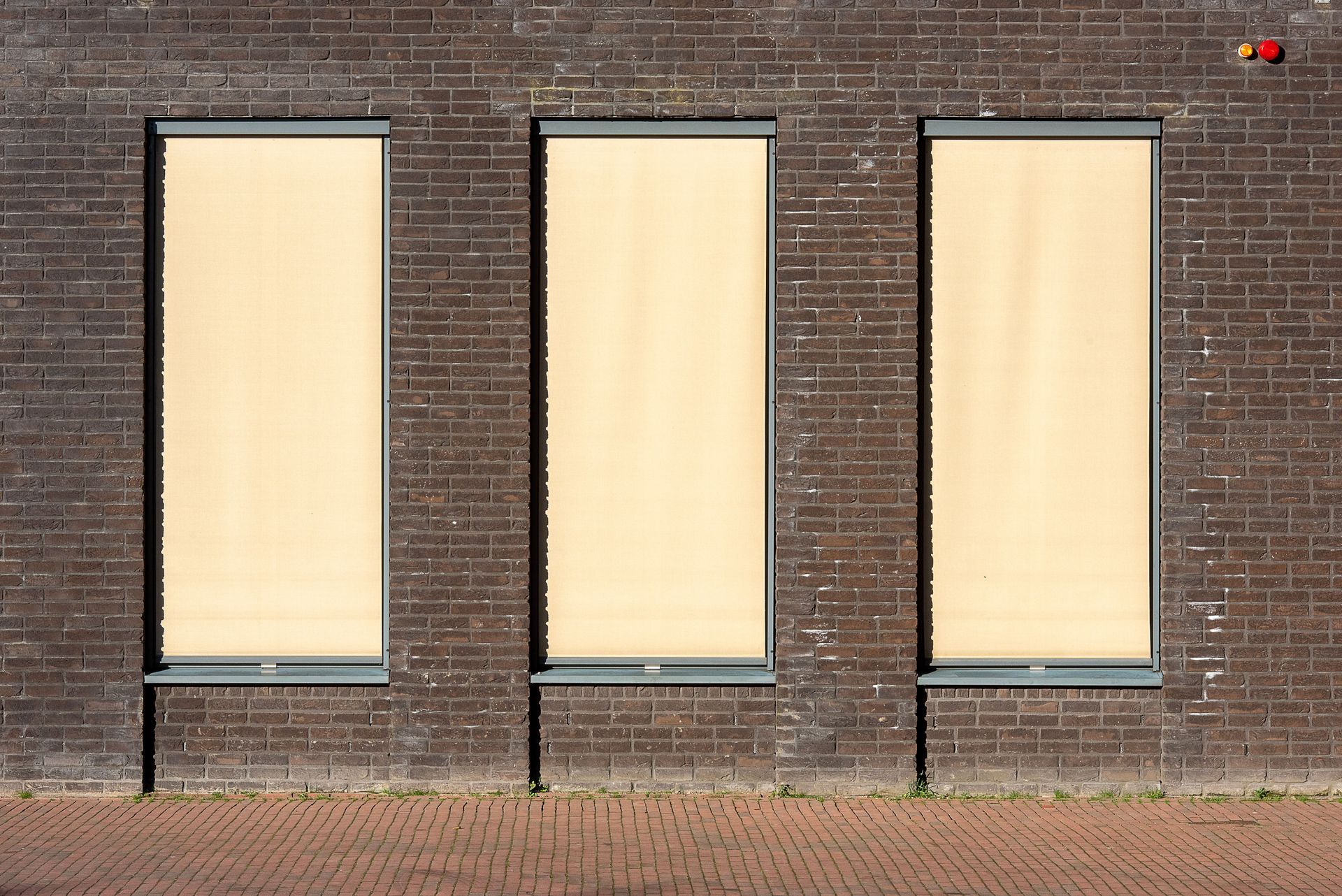 Three tall beige window shades on a brick wall above a paved sidewalk.