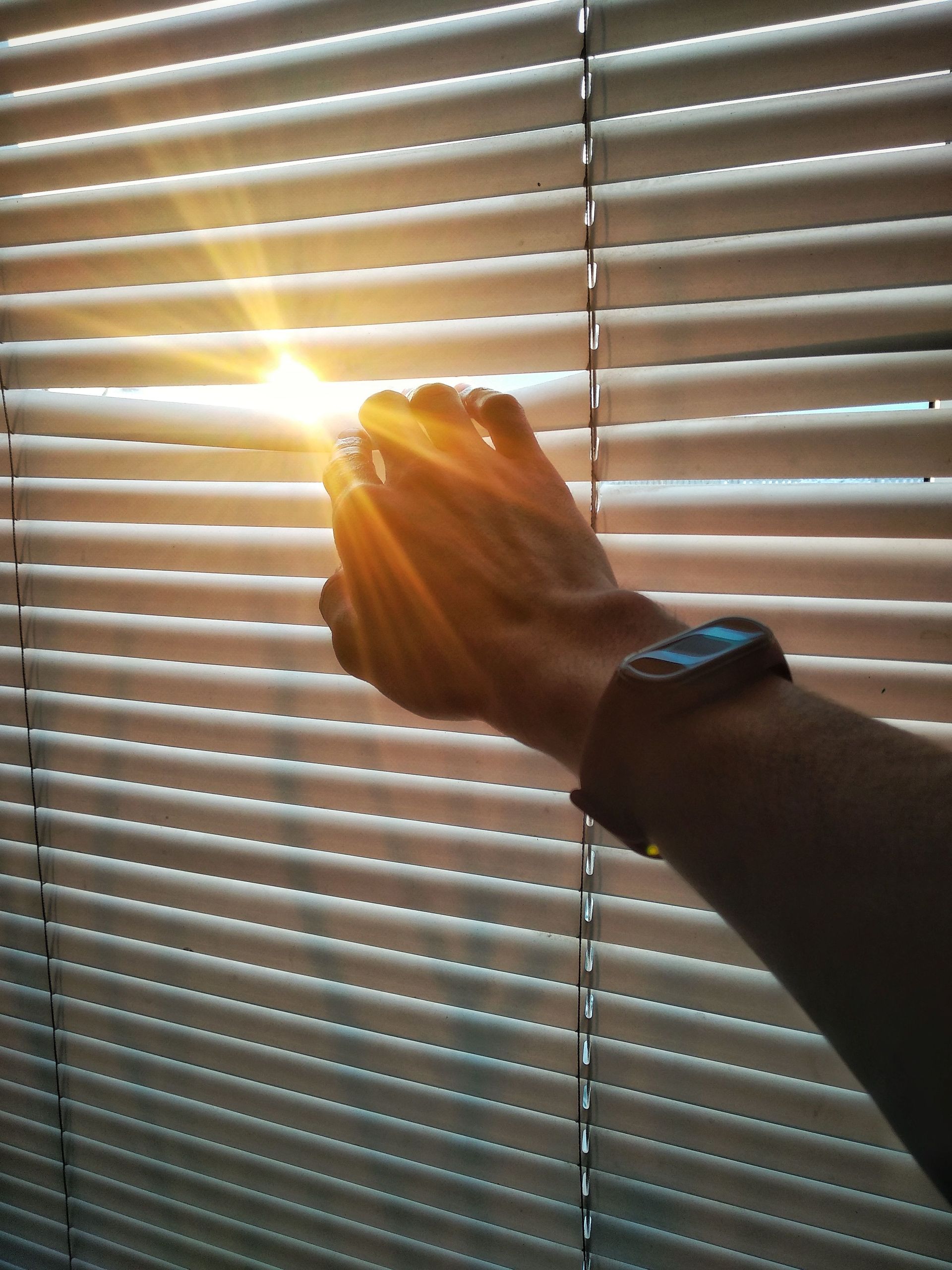 Hand reaching through window blinds toward bright sunlight, with a watch on the wrist
