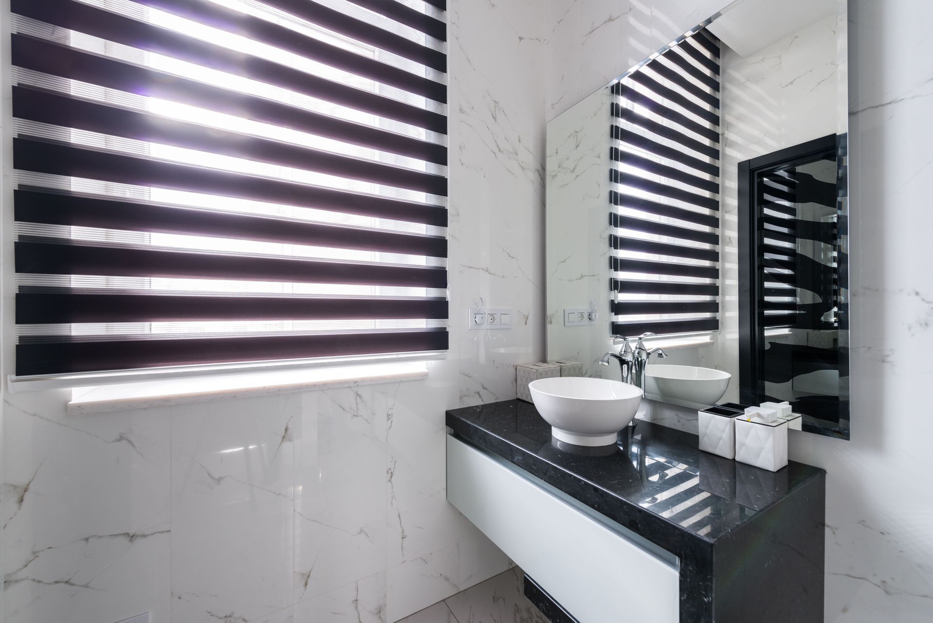 Modern bathroom vanity with black countertop, white vessel sink, mirror, and zebra blinds from two windows