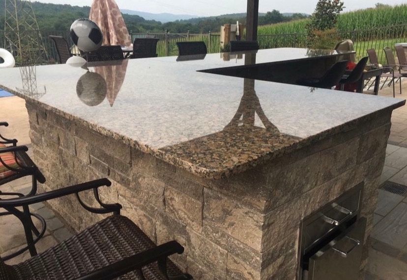 An outdoor stone kitchen island with a granite countertop, featuring a built-in drawer and bar seating on a patio.