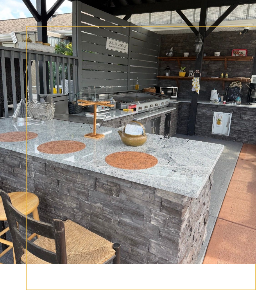 Outdoor kitchen with a granite-topped bar, stone base, wooden stools, and shelves against a dark tiled wall.