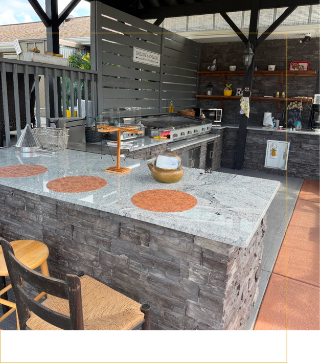 Outdoor kitchen with a stone-base granite island, wooden stools, and shelves against a dark grey stone backdrop.