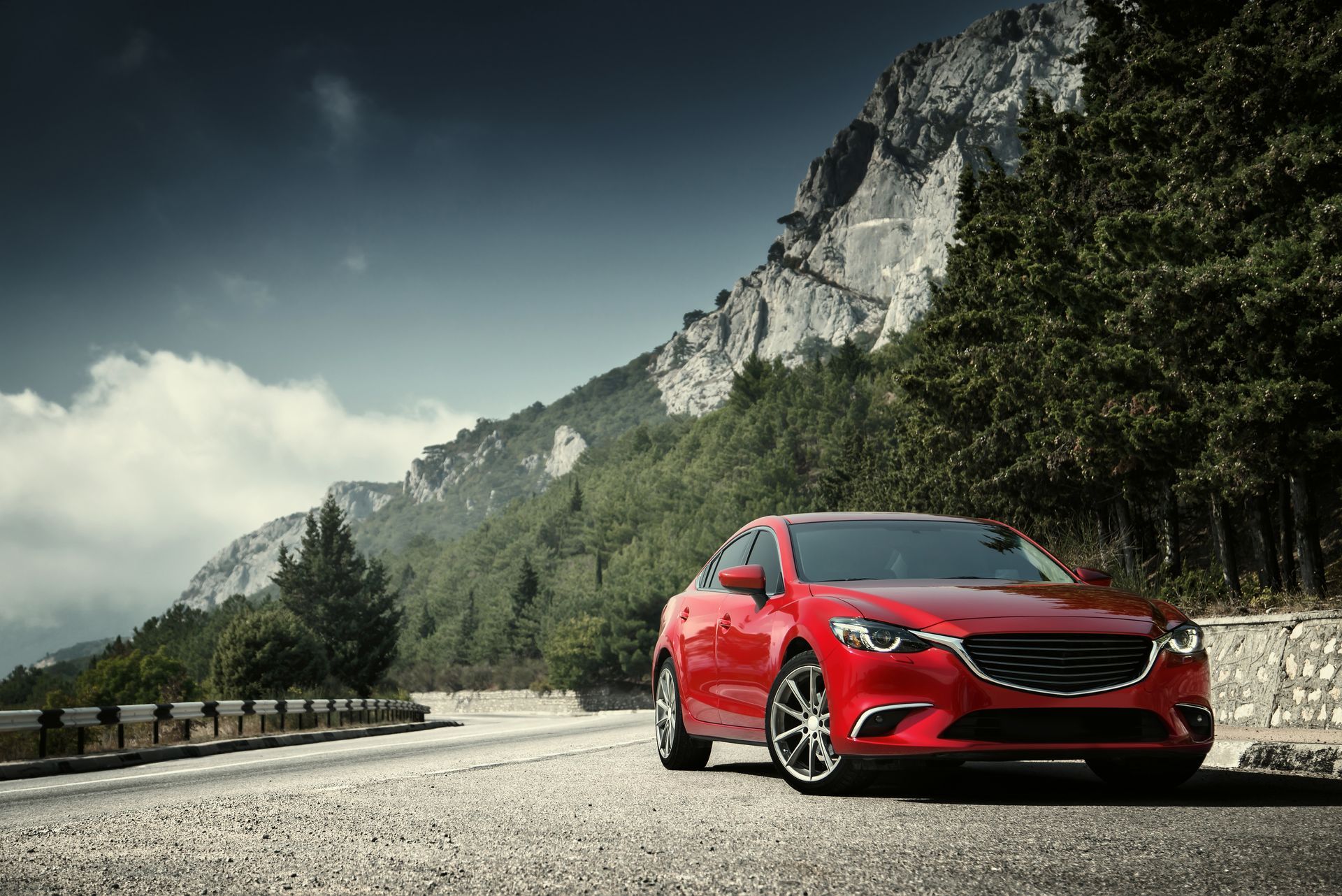 Red sports car on a mountain road with cliffs and trees under a cloudy sky | Cruz-N-Carz