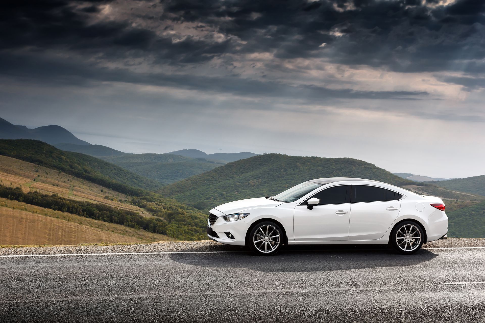 White sedan parked on a mountain road under a cloudy sky, with rolling hills in the background. | Cruz-N-Carz
