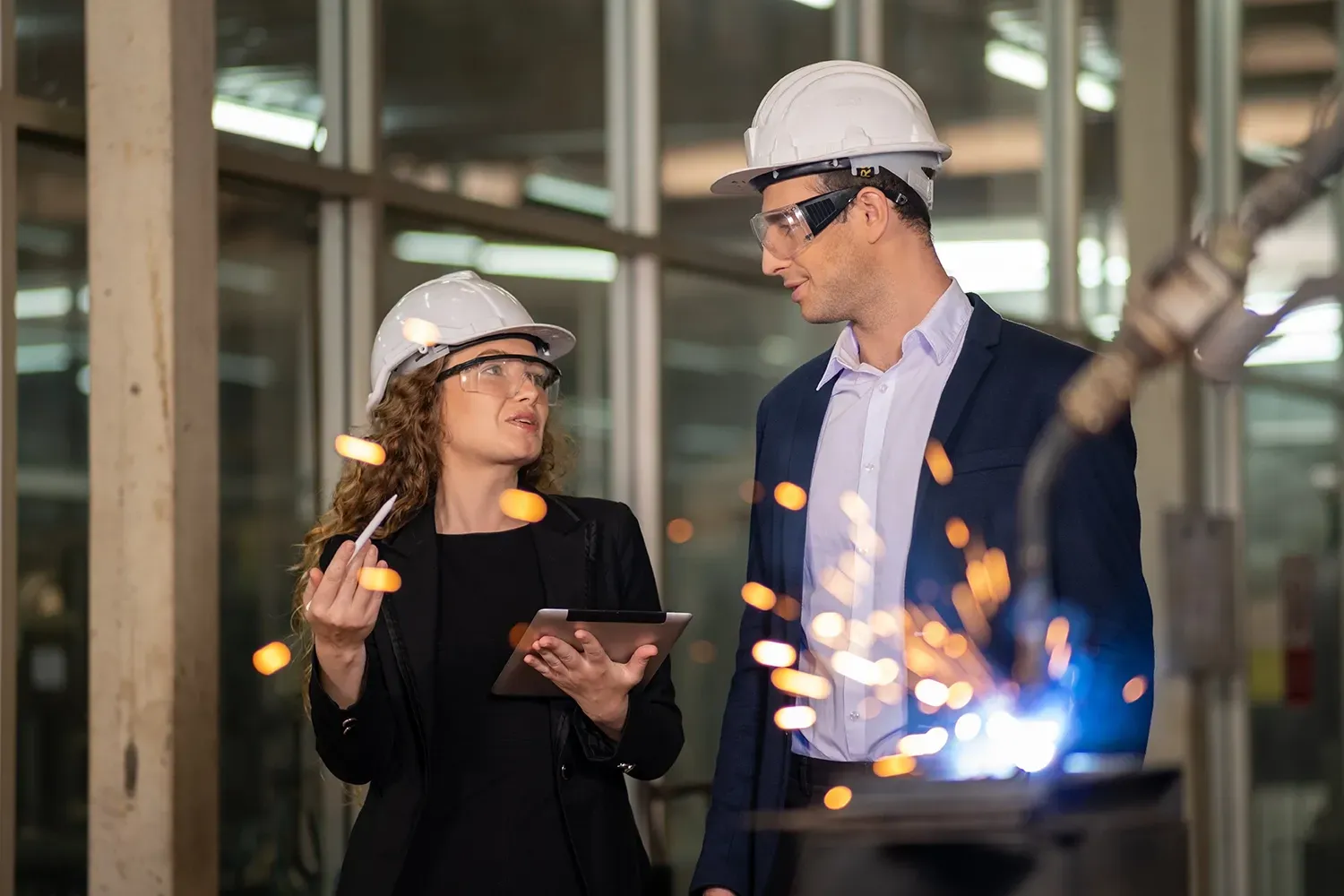 Two engineers in hard hats and safety glasses reviewing data on a tablet next to welding sparks duri