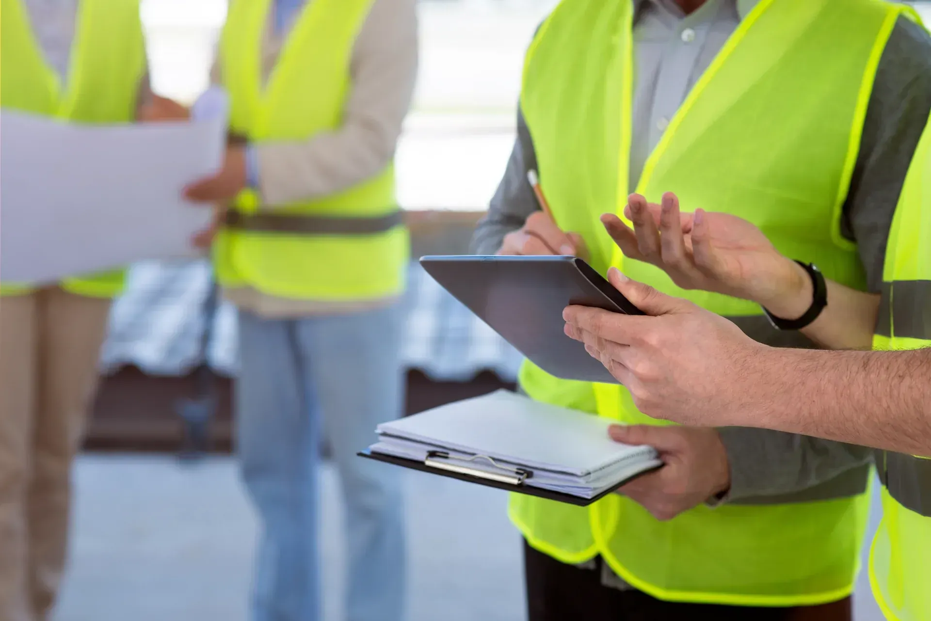 A close-up of an engineer in a high-visibility vest using a digital tablet and clipboard to review