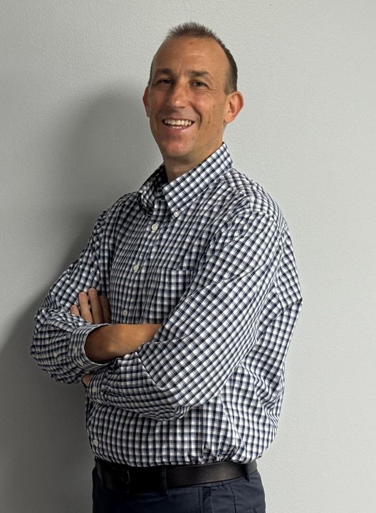 Smiling person in a checkered shirt standing with arms crossed against a plain wall