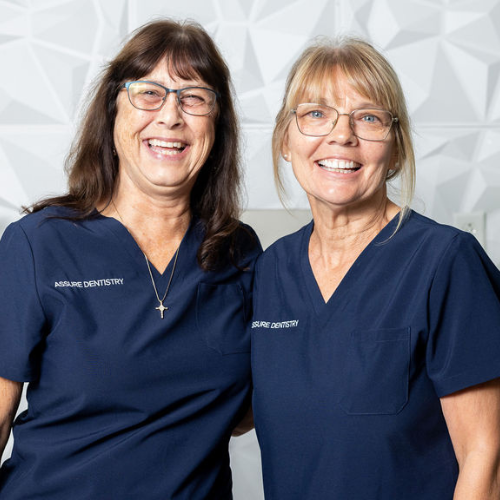Two women in blue scrubs smile, arms close, in front of a white wall.