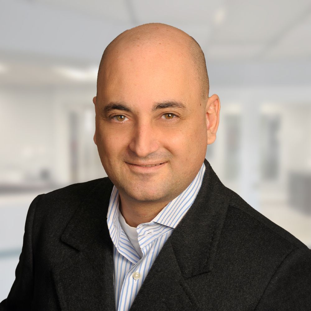 Bald man in black blazer and striped shirt smiles in a blurred office setting.