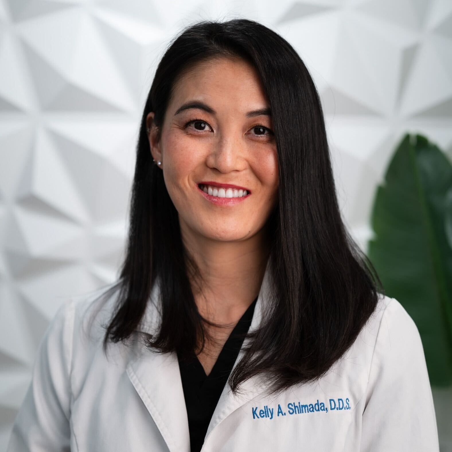A dentist in a white lab coat with her name embroidered on it, posing against a white geometric wall.