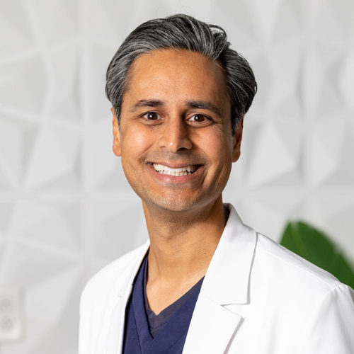 Man in a white coat smiles; light-skinned, graying hair, indoors against white textured wall.