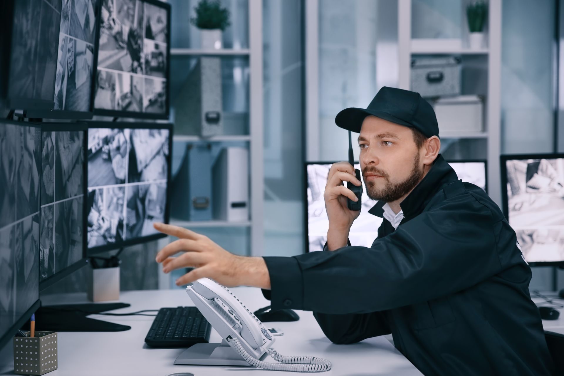Security guard using a two-way radio while monitoring surveillance screens in a control room.