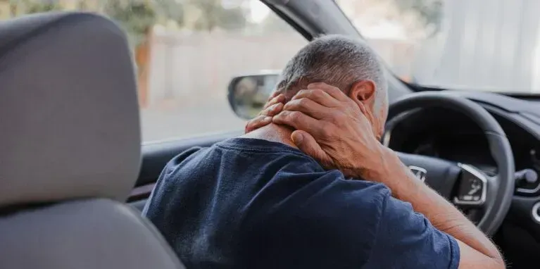 A man is sitting in the driver 's seat of a car holding his neck.