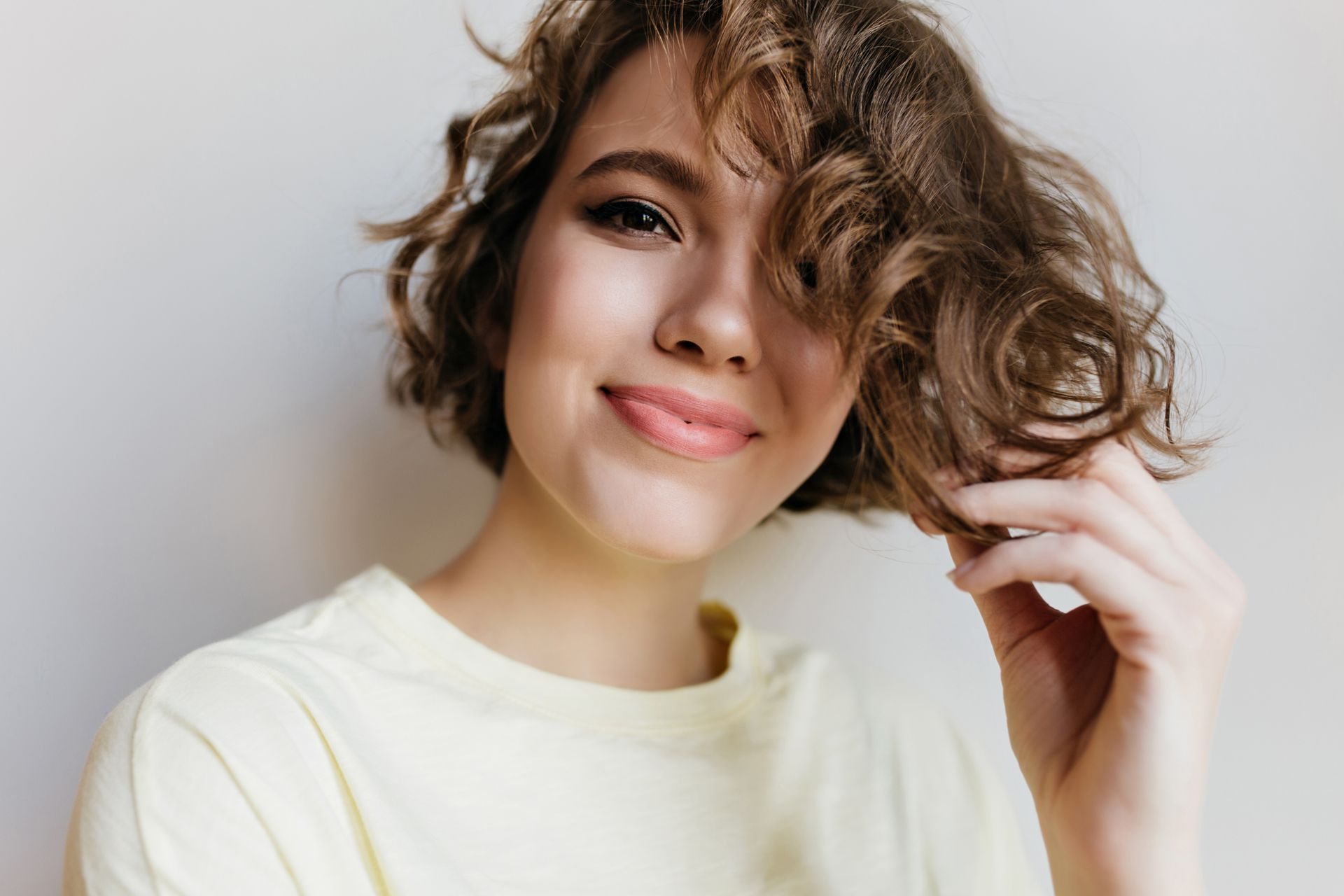 A woman with short curly hair is smiling and holding her hair.