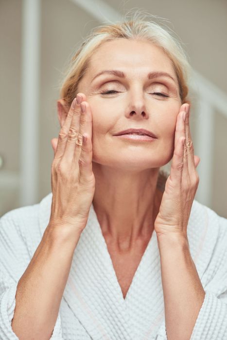 A woman in a bathrobe is applying lotion to her face.