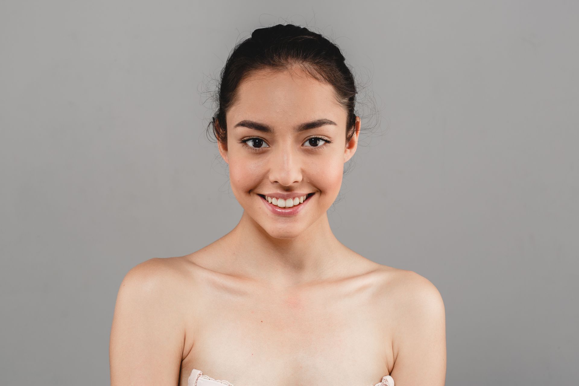 A woman without a shirt is smiling and looking at the camera over a gray background.