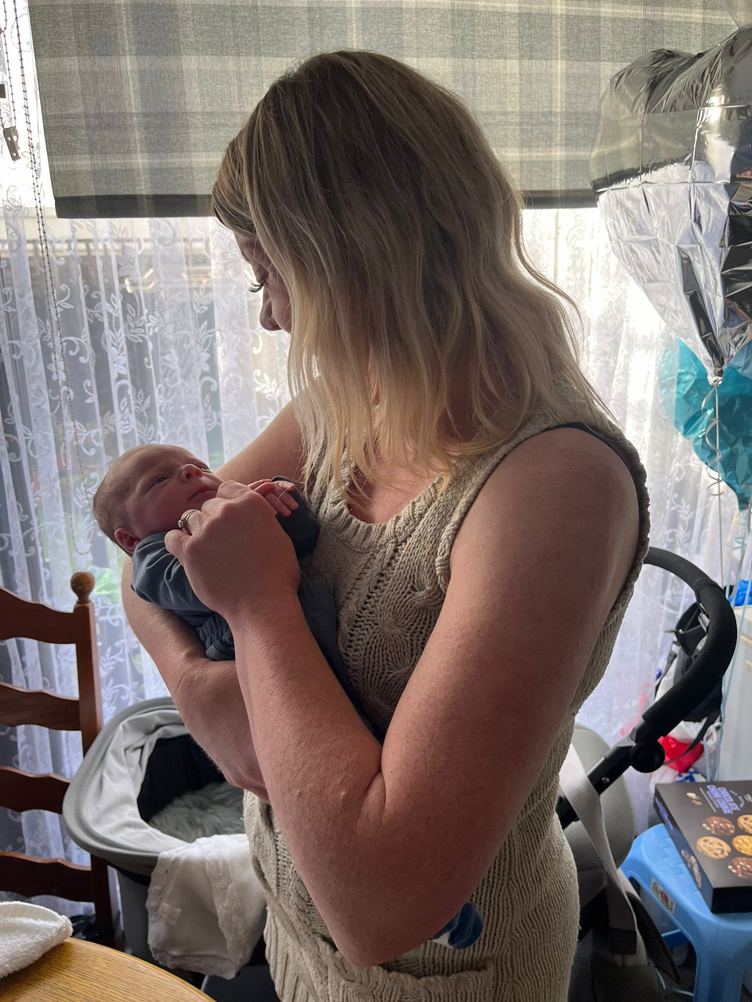 Woman in beige dress holding a newborn baby; they're inside near a window, with a bassinet and balloons in the background.