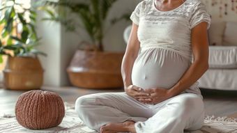 Pregnant person sitting cross-legged, hands on belly, indoors with plants and neutral decor.