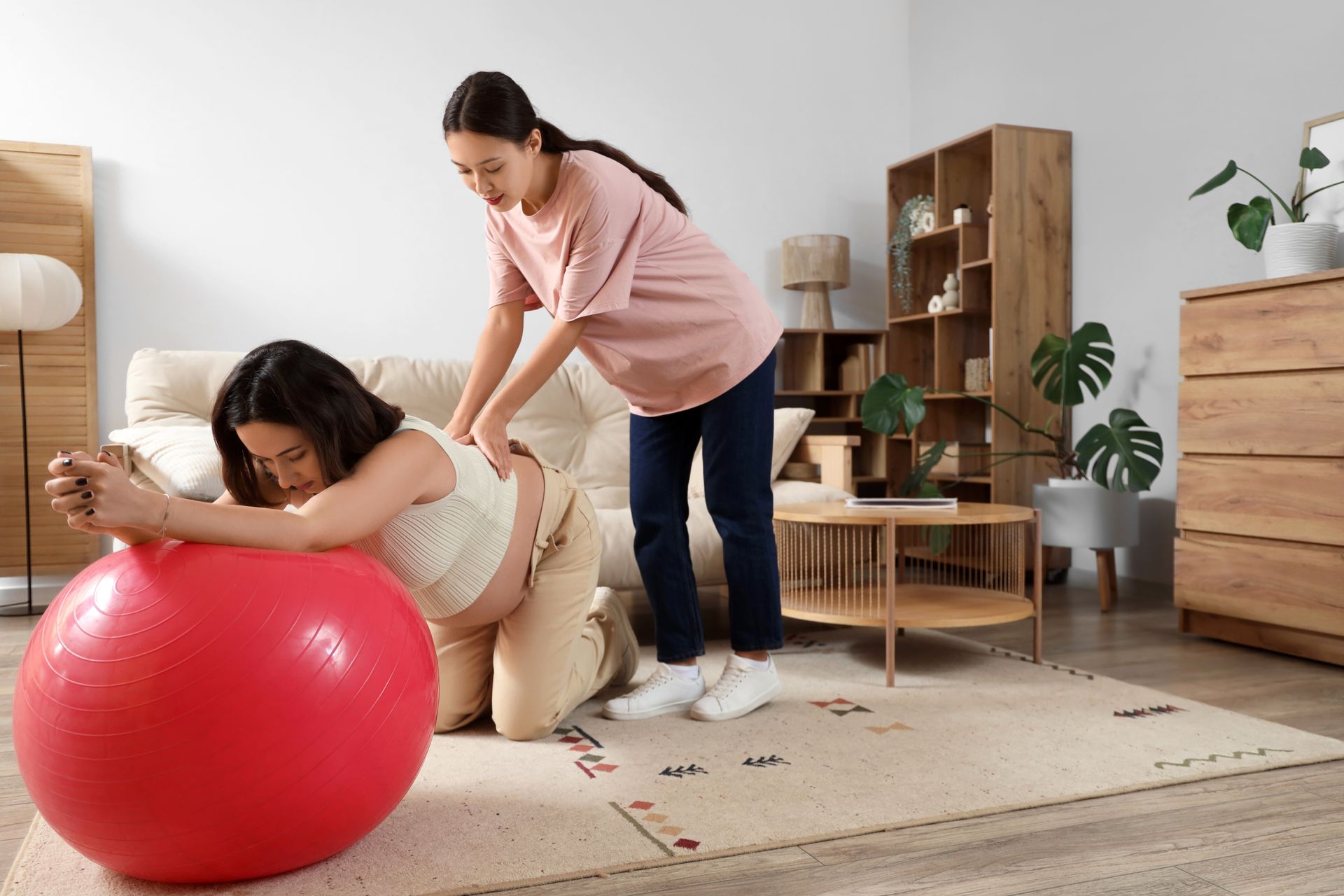 Pregnant woman using exercise ball, another woman supporting her back, home interior.