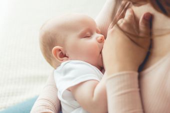 Baby nursing from a person. Baby is wearing a white onesie, eyes closed. Person's arm is wrapped around baby.