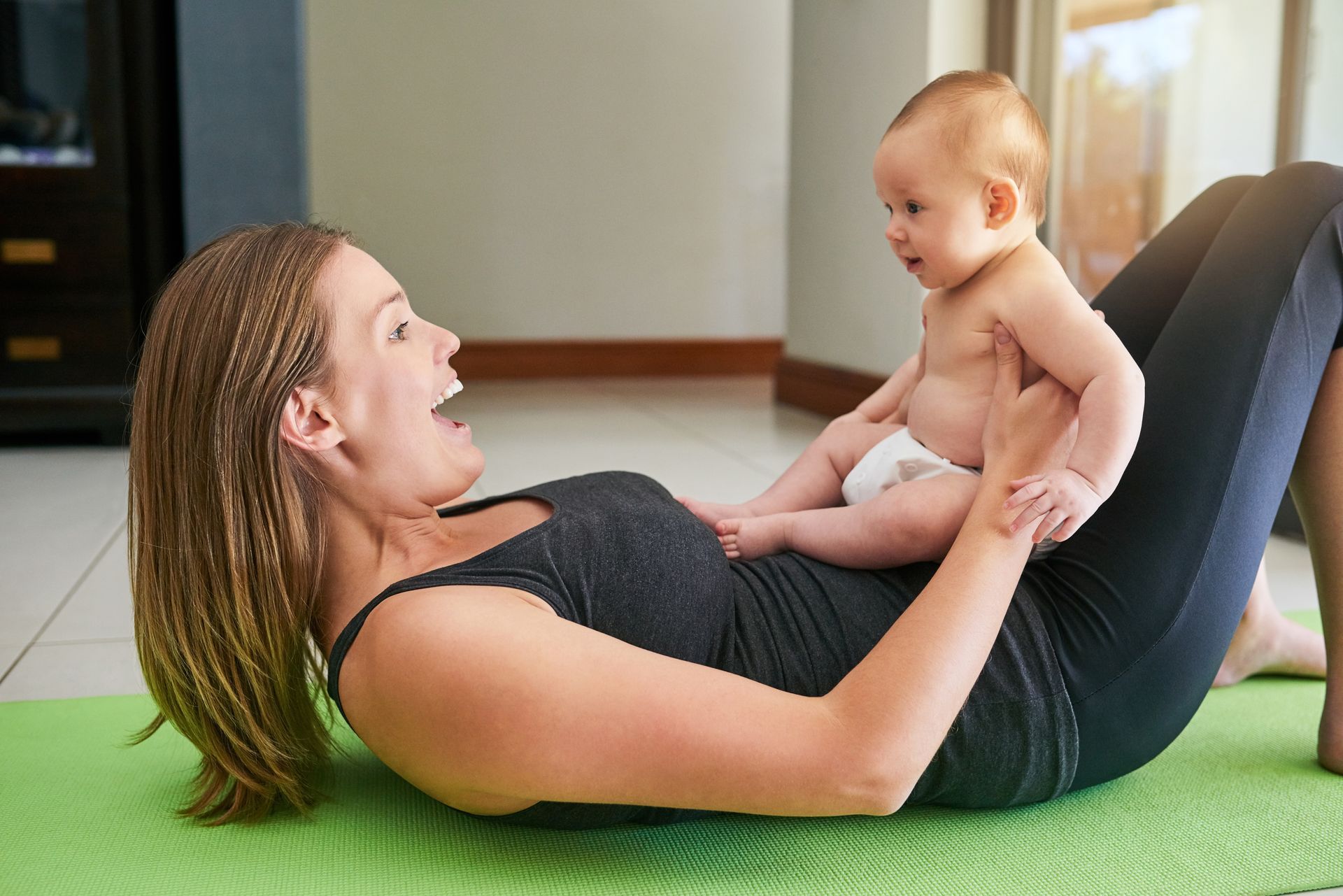 Woman exercising on a mat with a baby; woman smiling, baby looking up. Indoors.