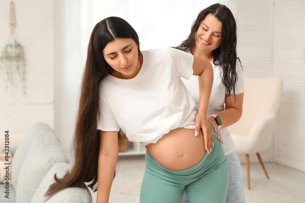 Pregnant person, wearing green pants, bent over; another person supports them. Inside.