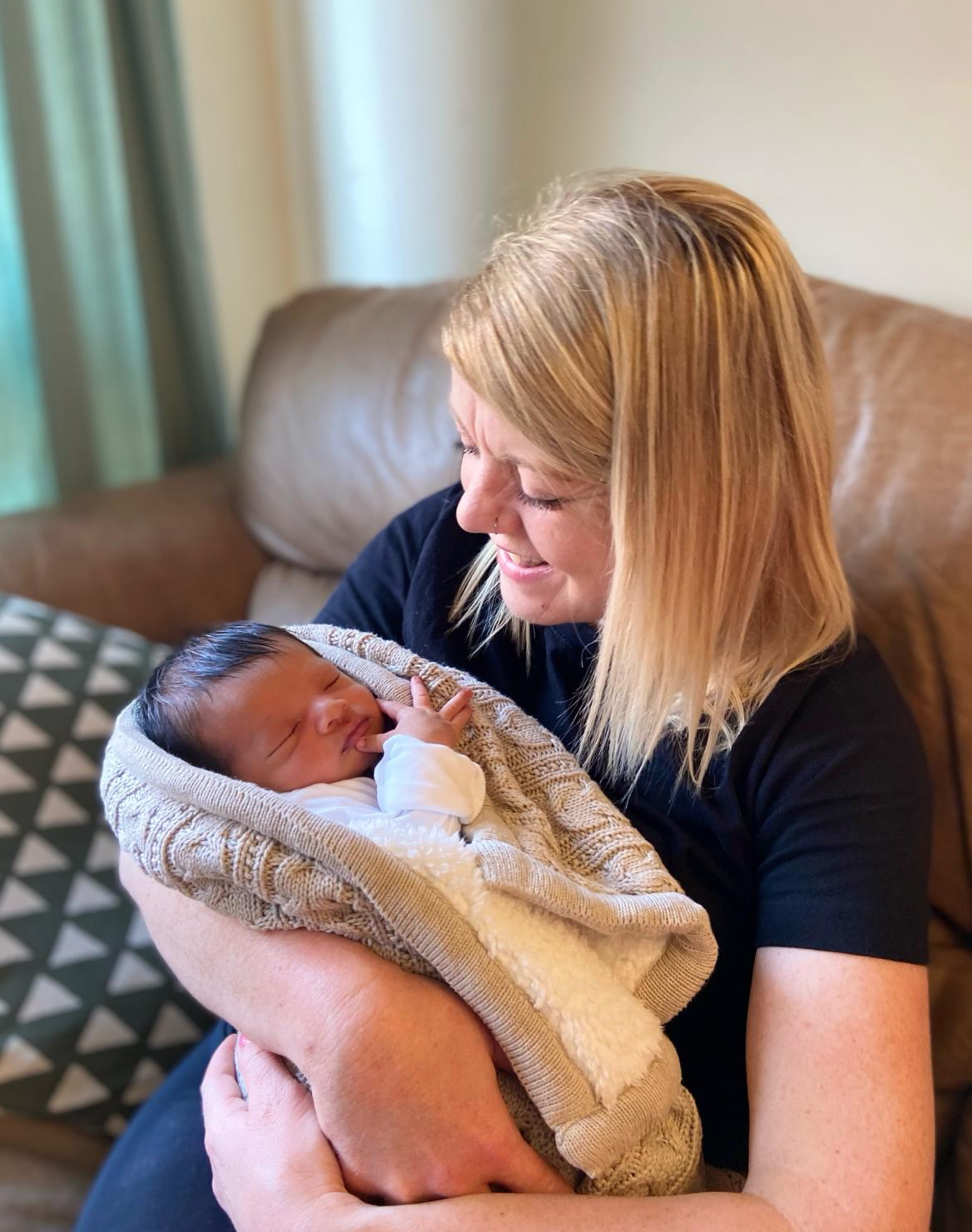 Woman holding a swaddled newborn, smiling. Tan blanket, soft lighting, seated indoors.