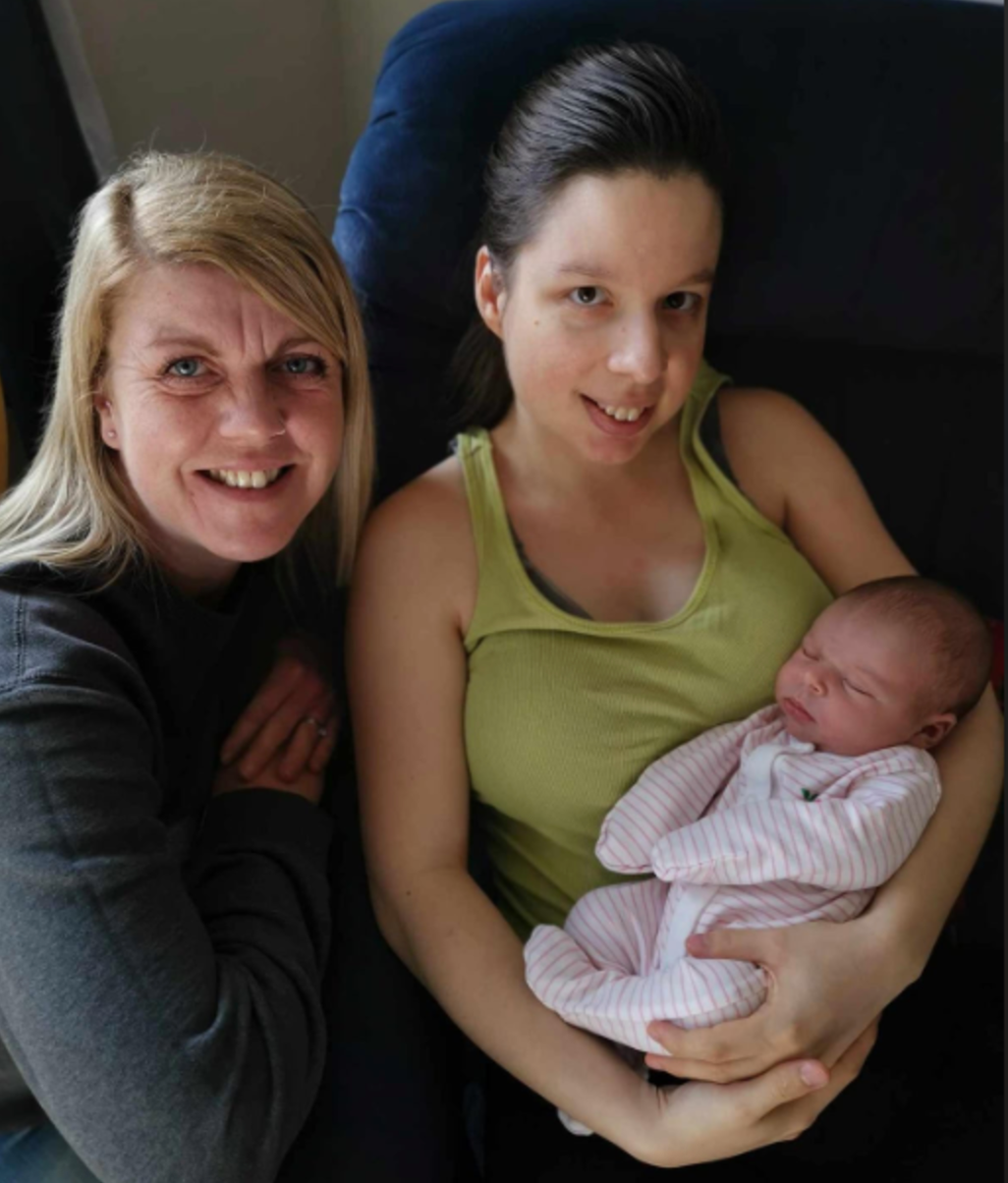 Two women, one holding a newborn baby. All three are smiling. Indoor setting, natural light.