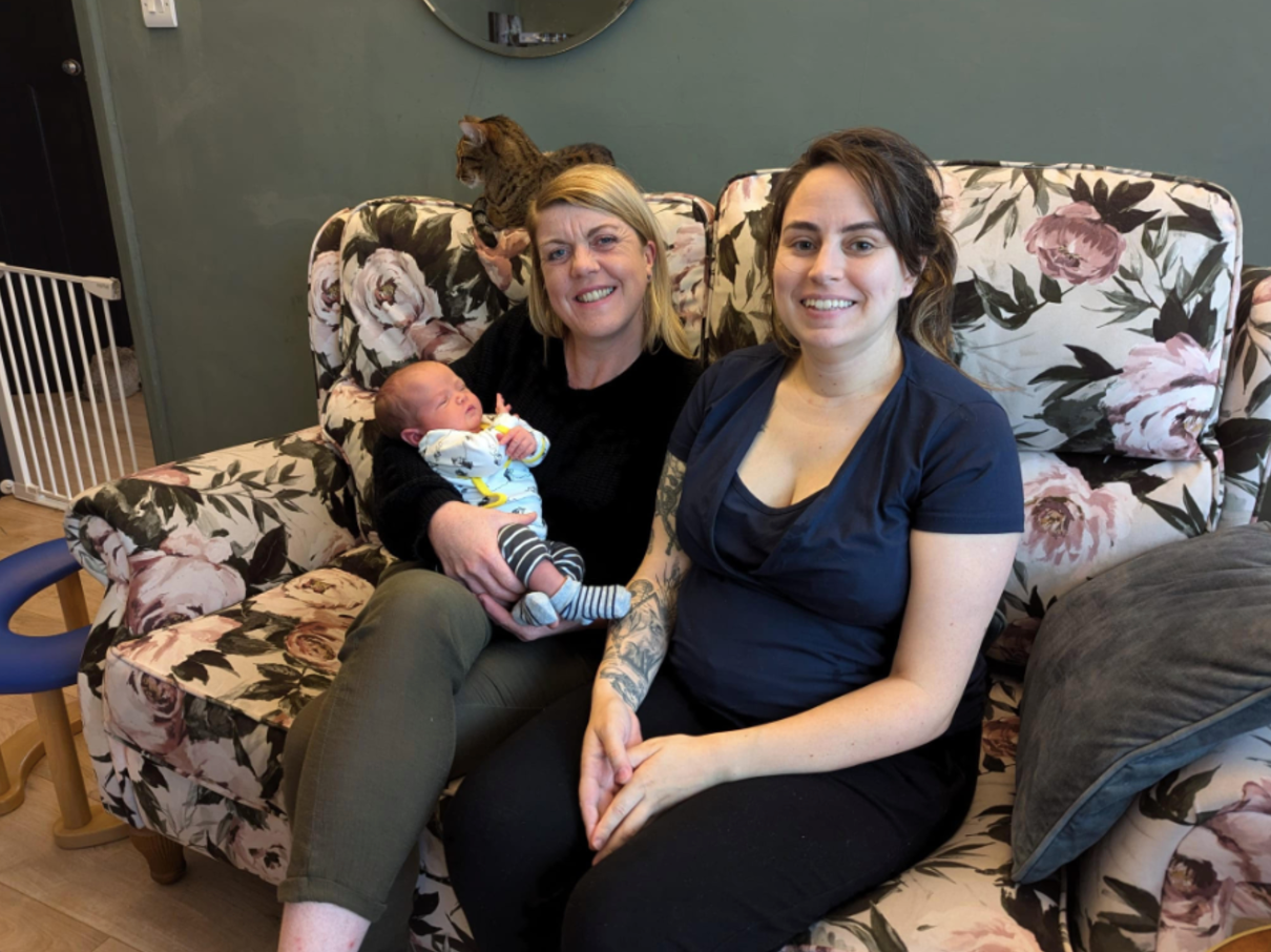 Woman holding baby, sitting with another woman on floral sofa. Cat perched above them.