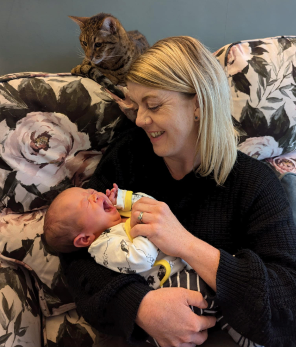 Woman smiling at a crying baby in her arms. A cat watches from behind on floral sofa.