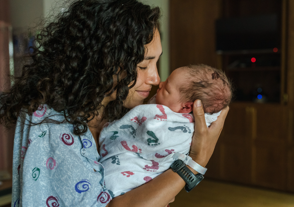 Woman holding and gazing at a newborn baby, both touching noses; hospital room setting.