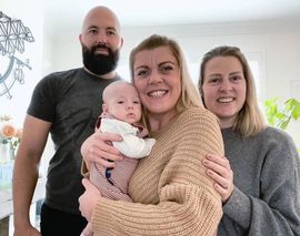 Family of four: man, woman holding baby, and another woman smiling indoors.