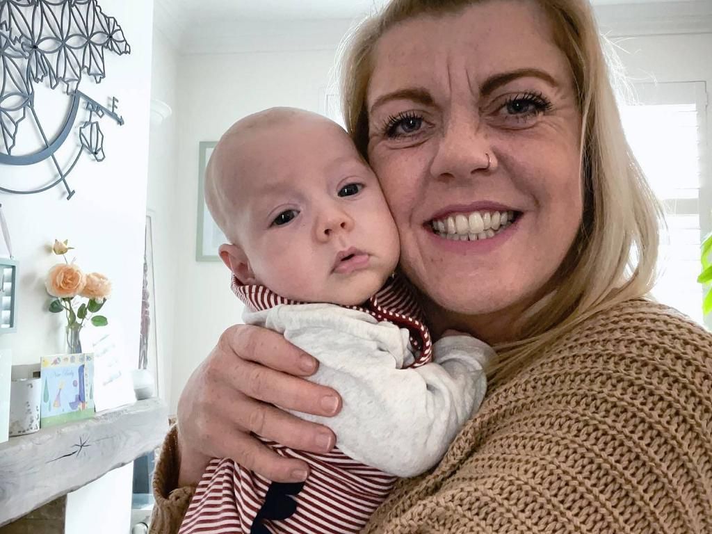 Woman holding a baby, both looking at the camera. Woman is smiling; the baby has a neutral expression. Indoors.