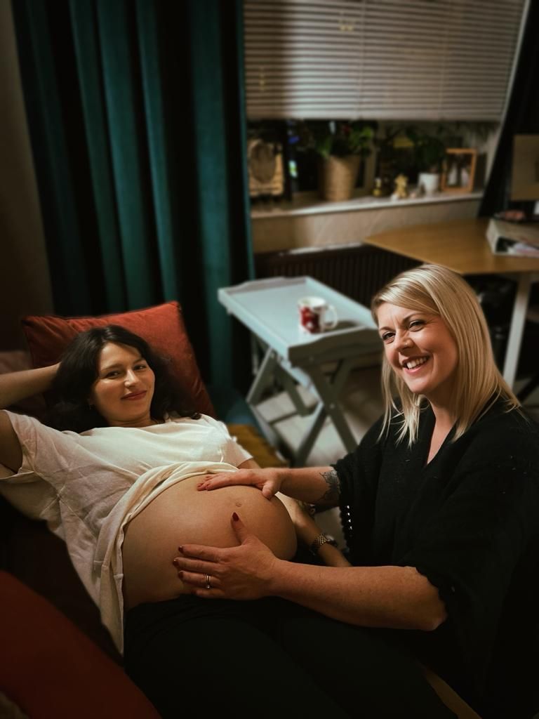 Pregnant person and blonde woman smiling, touching the pregnant belly indoors with curtain and table.