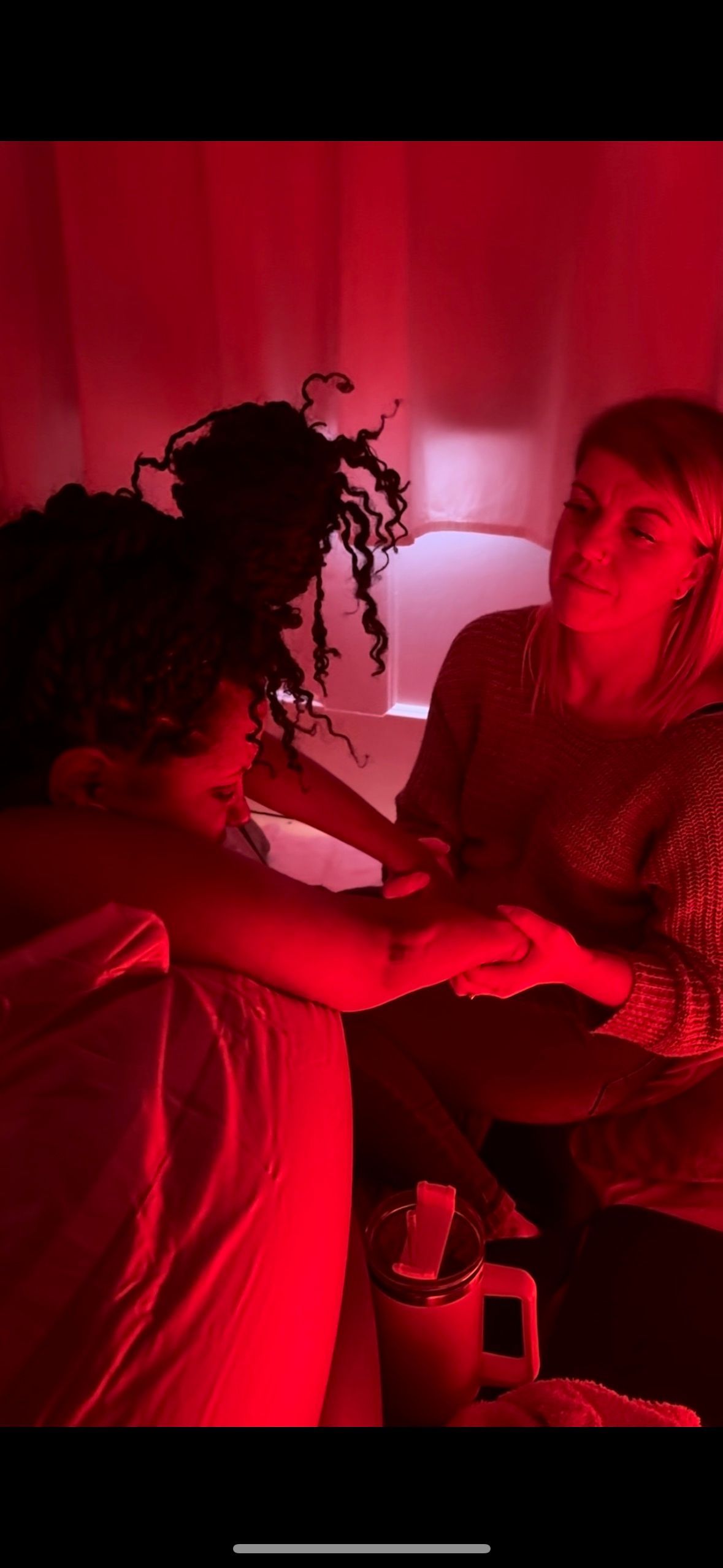 Two people touching hands in a red-lit room. One with dark curly hair.