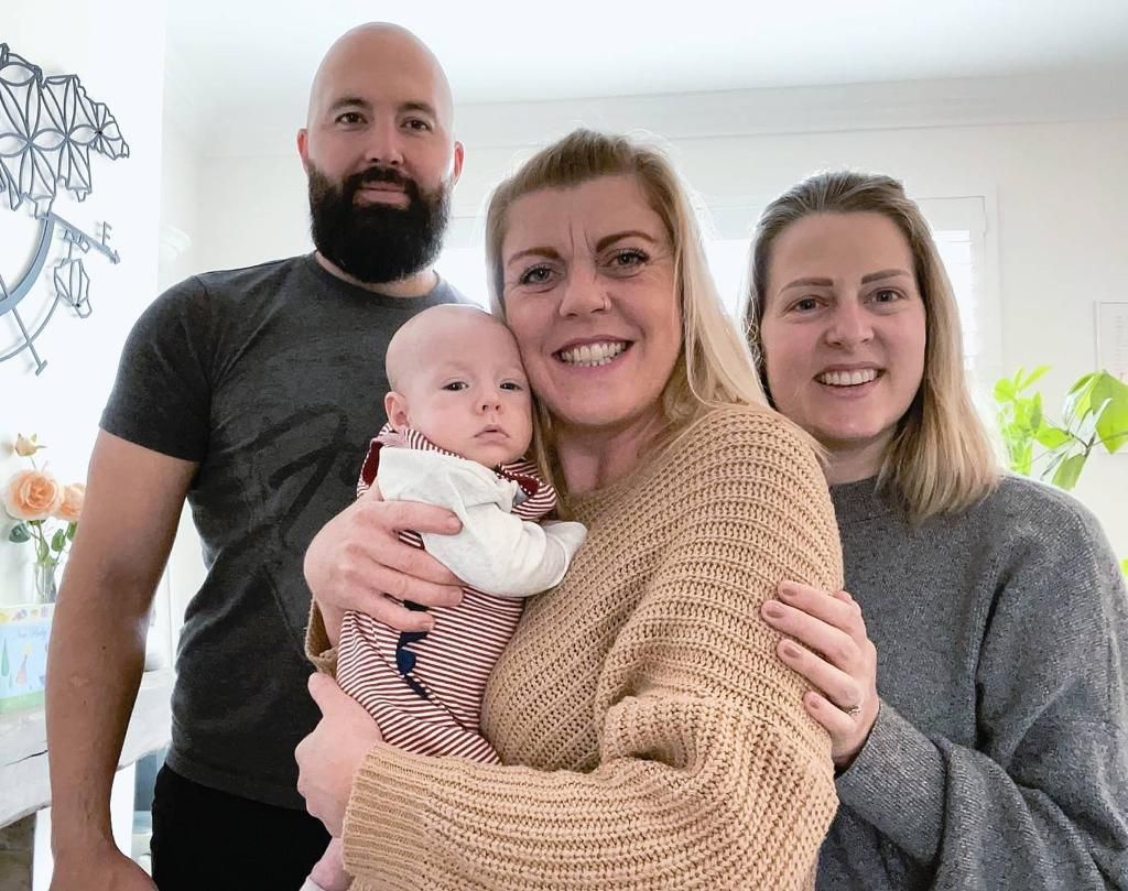 Family of four smiling, holding baby. Two women and a man in a home setting.