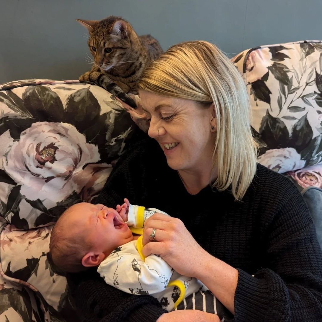 Woman holding crying baby, cat watches from floral couch.