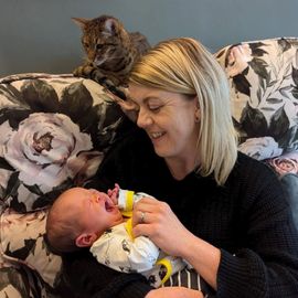 Woman holding crying baby, cat watches from floral couch.