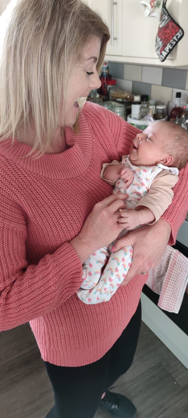 Woman in pink sweater holds a smiling baby in a kitchen. The baby wears floral pajamas.