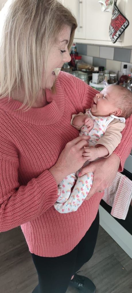 Woman in pink sweater holds a smiling baby in a kitchen. The baby wears floral pajamas.