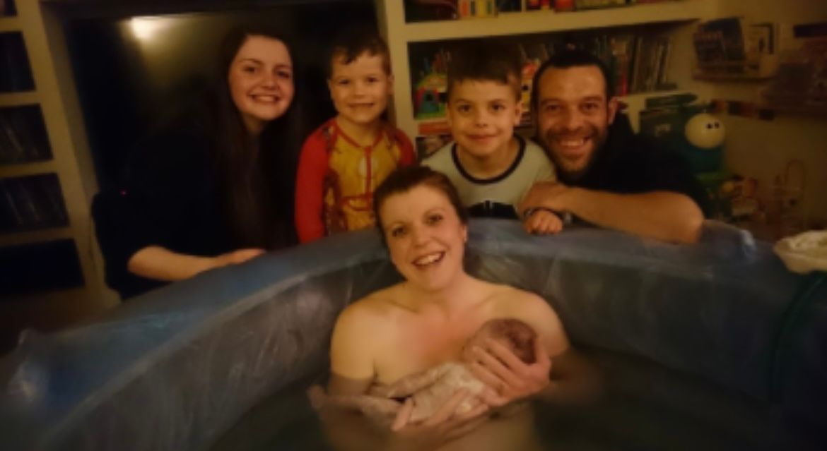 Family gathers around a woman holding a newborn in a birthing pool. Smiling faces.